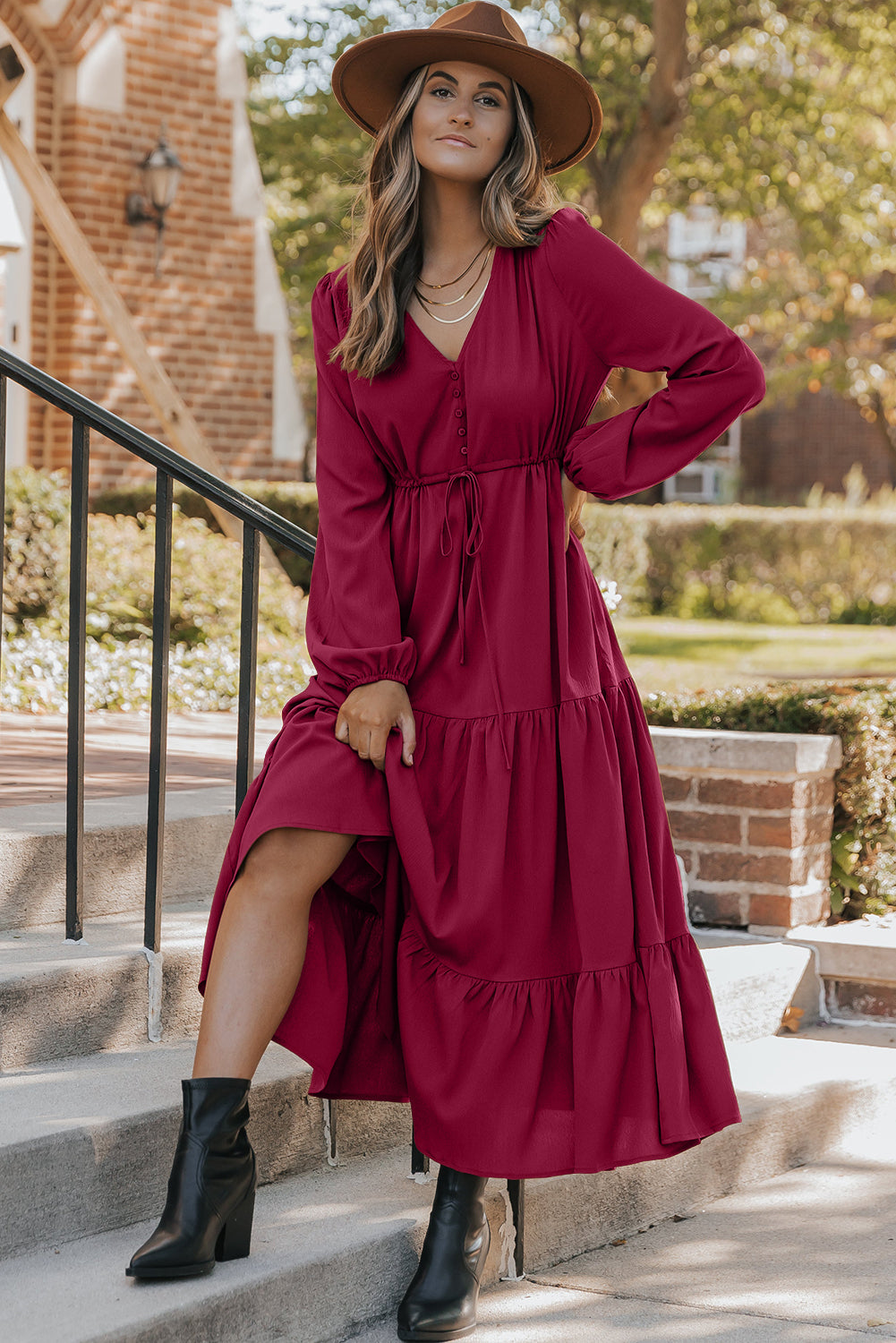Woman in a long-sleeve burgundy maxi dress, brown hat, and black ankle boots posing outdoors on steps