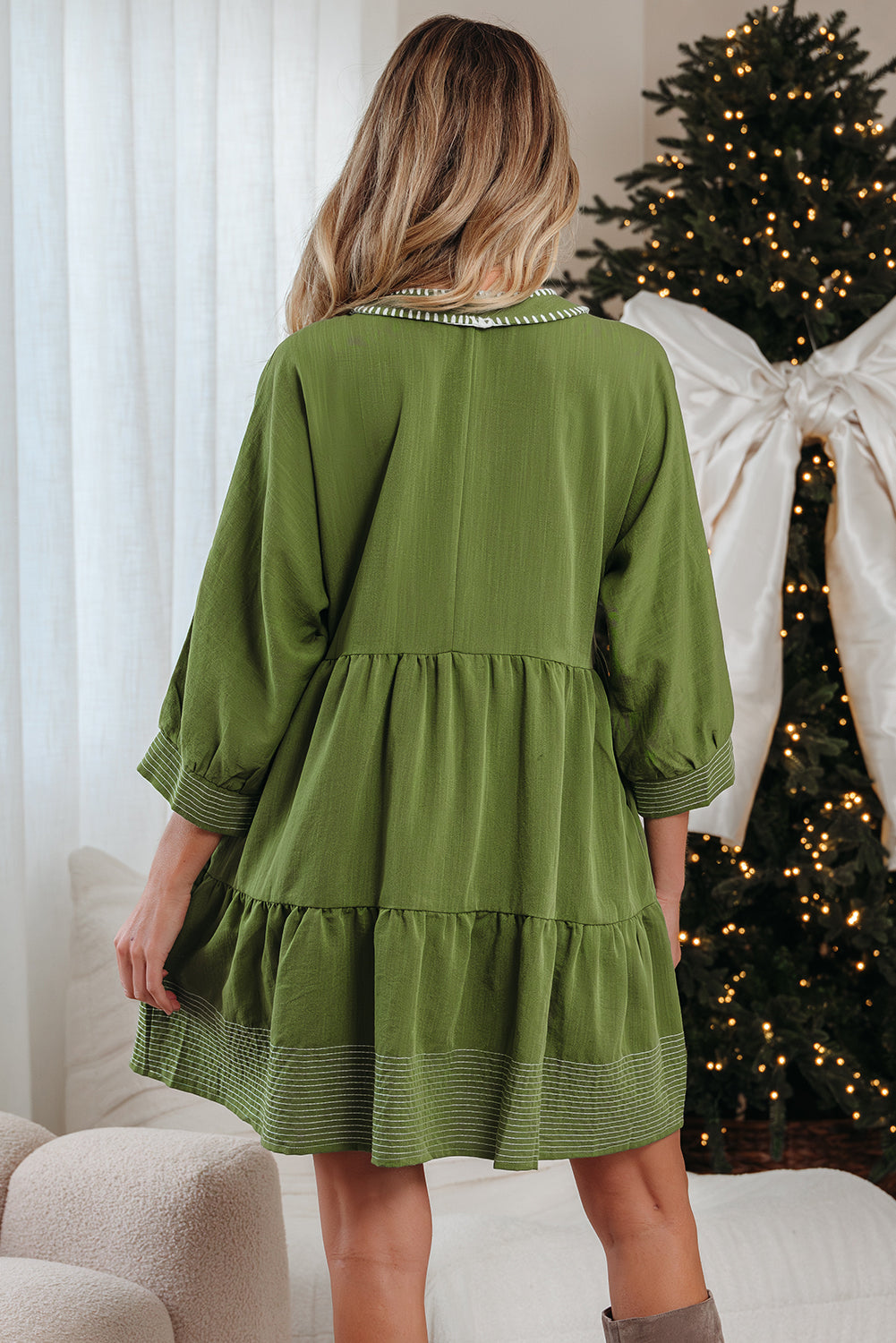 Woman in green tiered dress with striped trim, standing indoors by a decorated Christmas tree