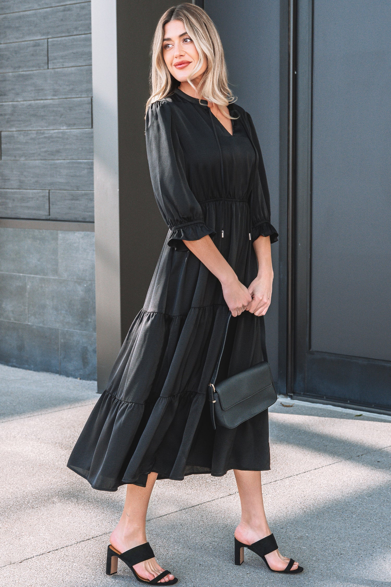 Woman wearing a black midi dress with ruffled sleeves, black sandals, and a black clutch bag