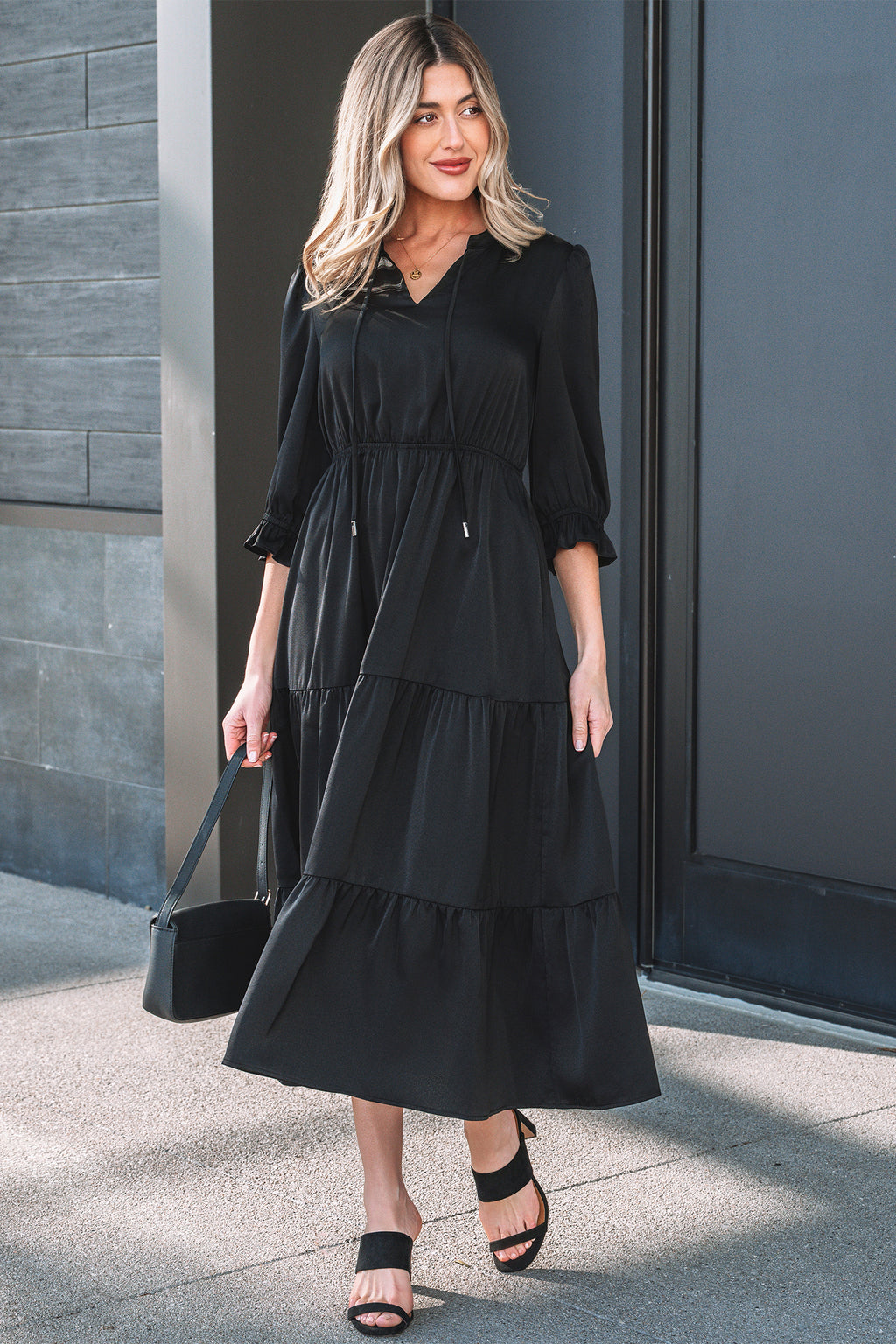 Woman in a black midi tiered dress with puff sleeves, black sandals, and a black handbag