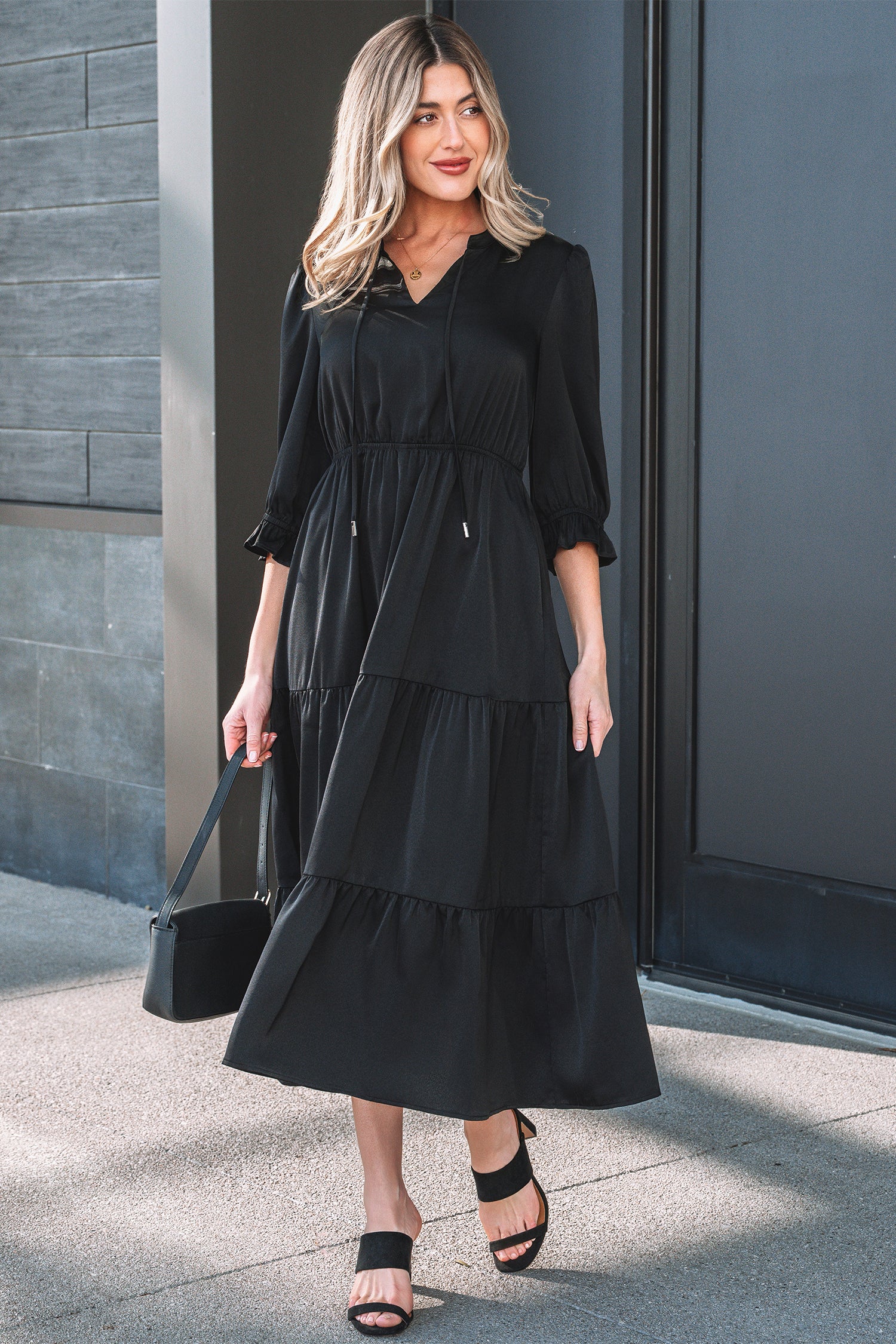 Woman in a black midi tiered dress with puff sleeves, black sandals, and a black handbag