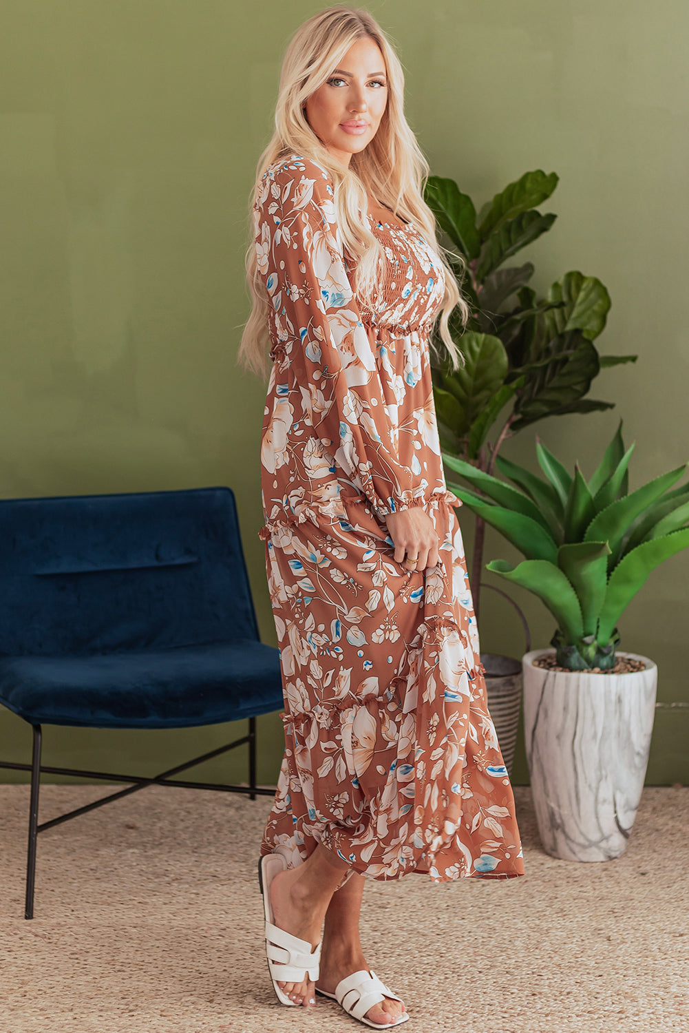 Woman in long floral brown dress and white sandals standing indoors near plants and chair