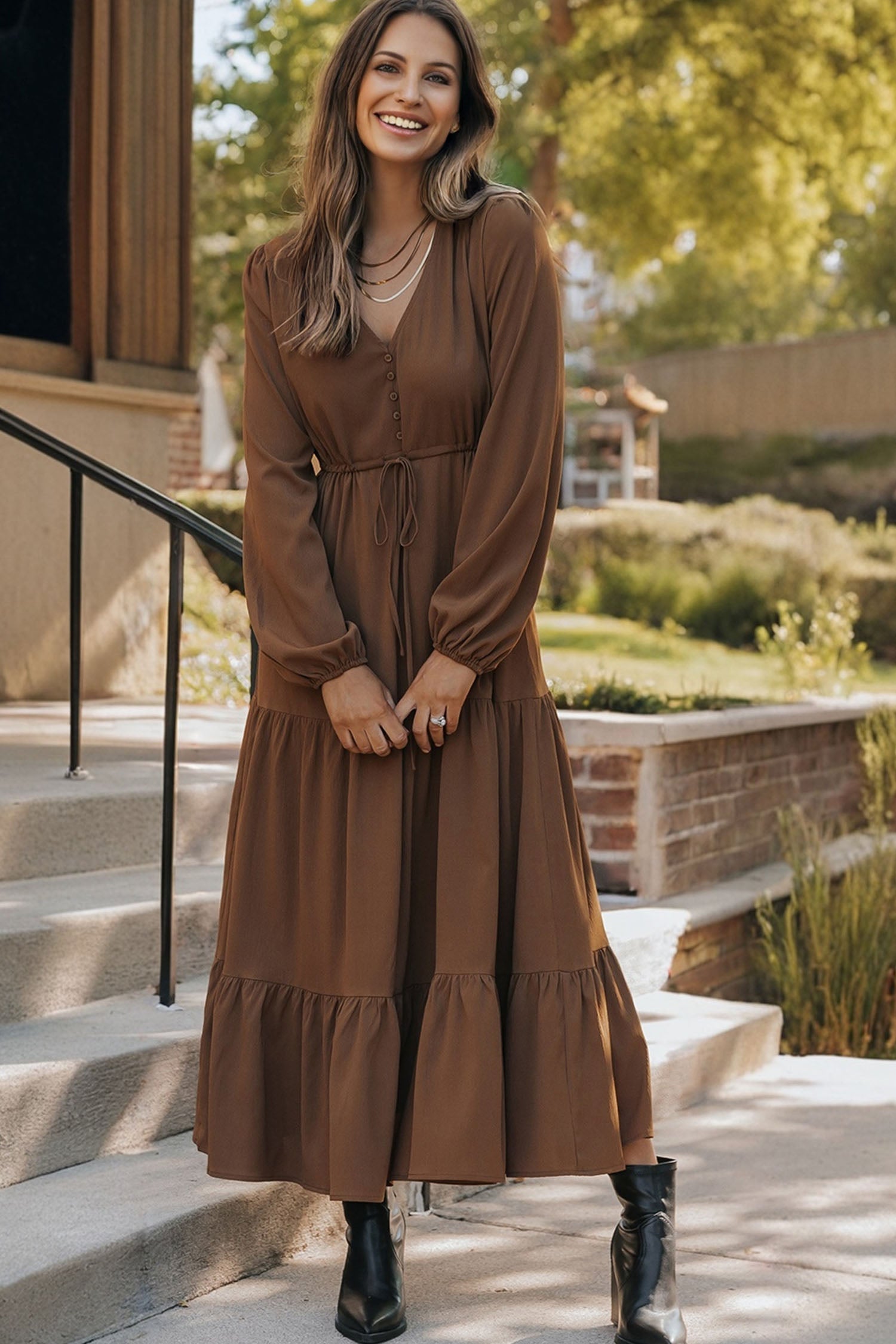 Woman in a brown long-sleeve maxi dress and black boots, smiling outdoors in sunlight.