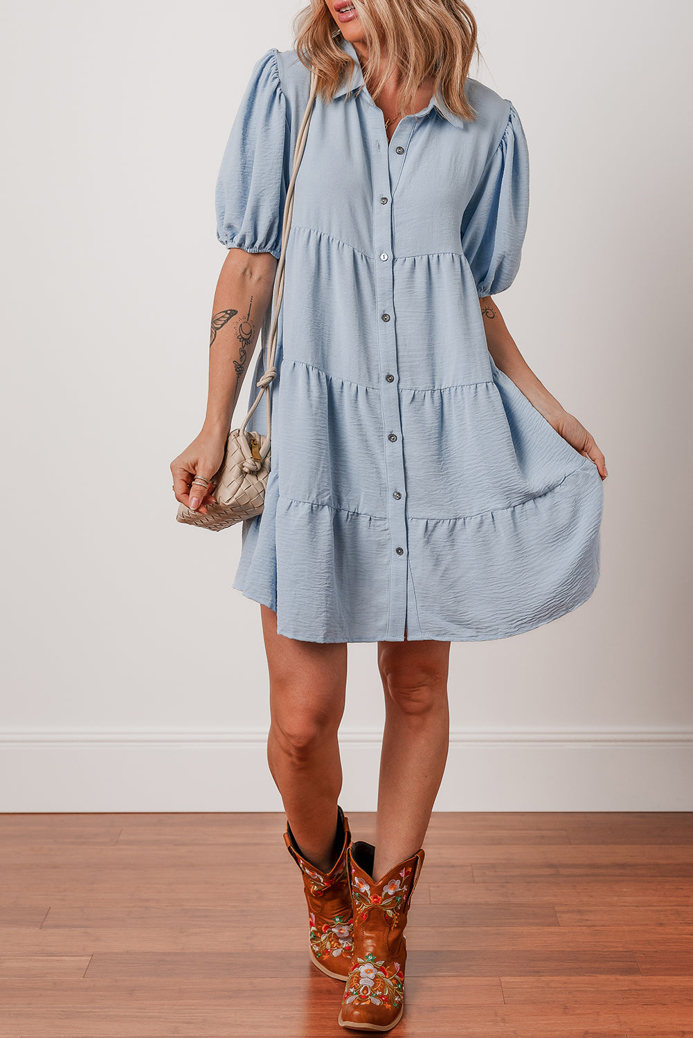 Woman wearing light blue short button-up dress with puff sleeves, floral cowboy boots, and a woven purse