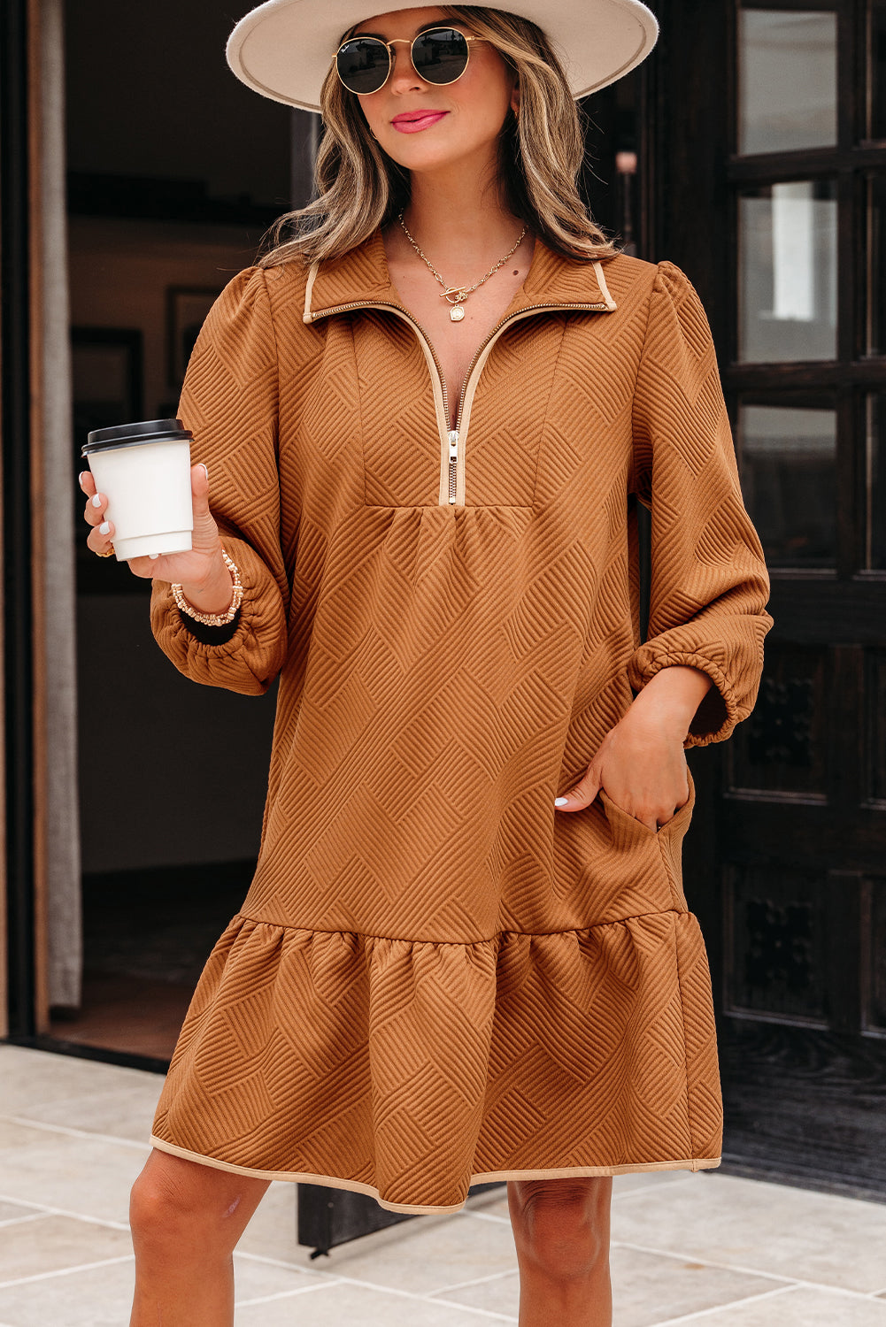 Woman wearing a brown textured long-sleeve dress with zipper neckline, white hat, and sunglasses, holding a coffee cup