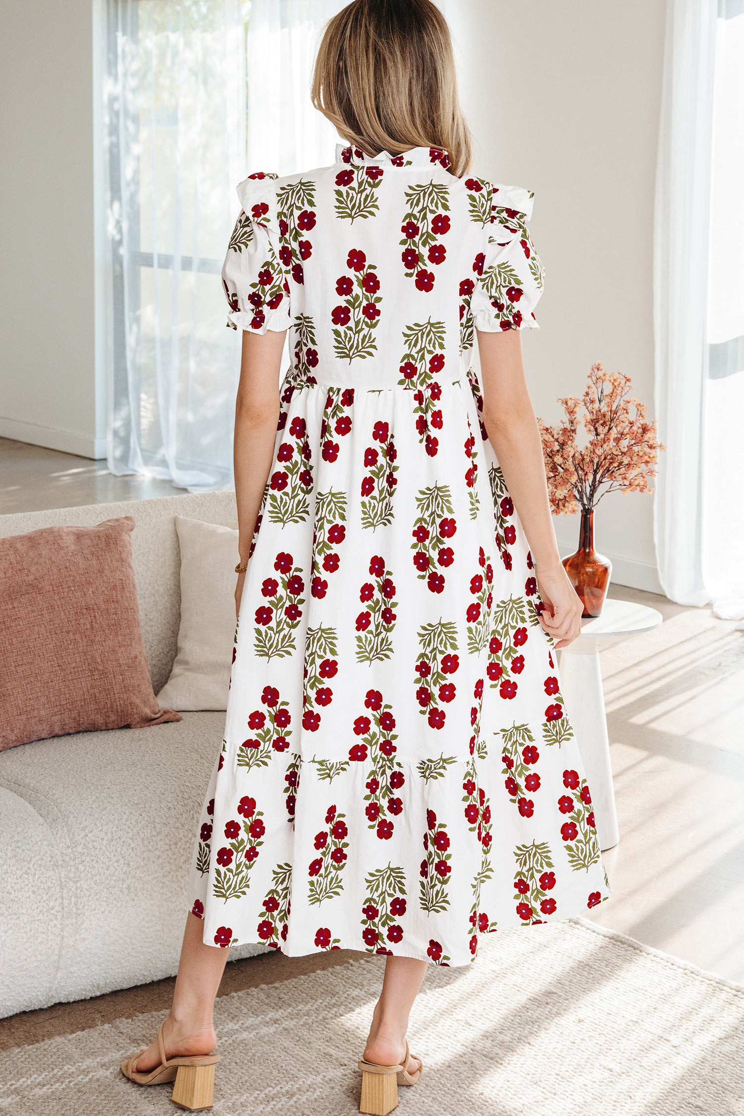 Woman in a white floral midi dress with red and green flower print, standing indoors