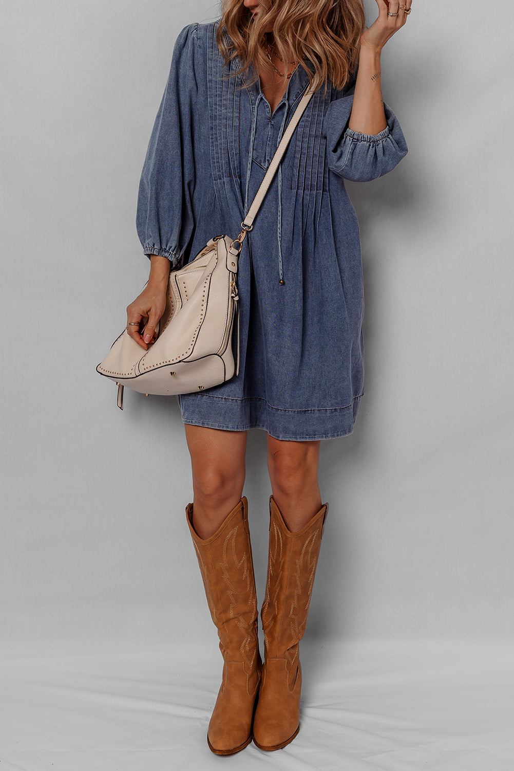 Woman wearing denim dress, tan knee-high boots, and beige leather shoulder bag.