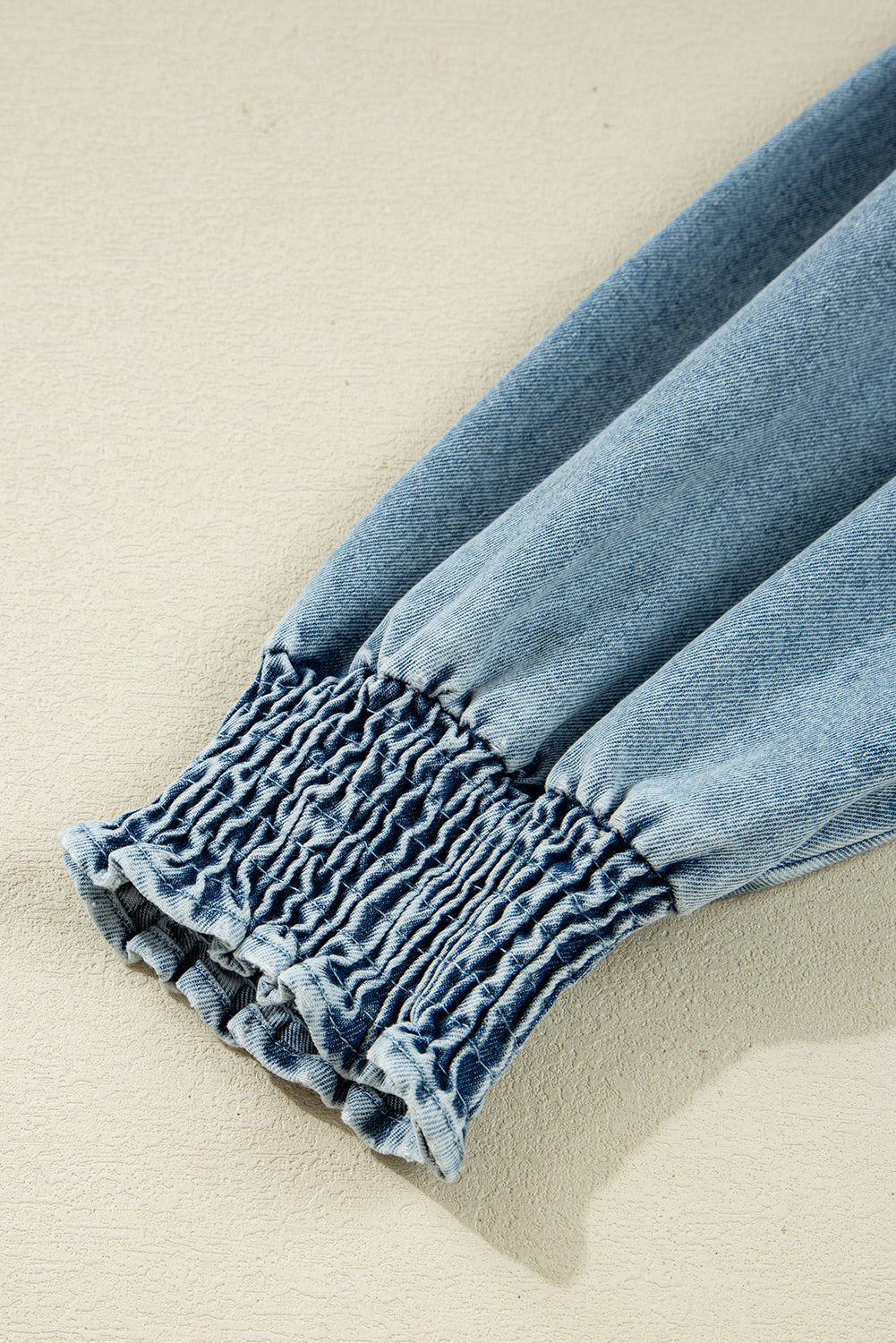 Close-up of a blue denim sleeve with elastic smocked cuff on a neutral background