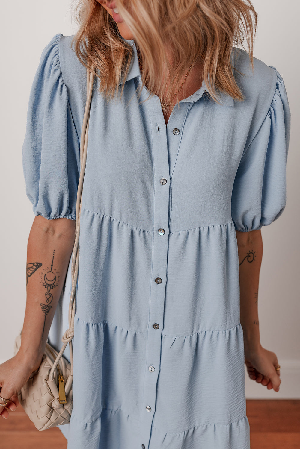 Woman wearing a light blue button-down dress with puff sleeves, holding a woven beige purse