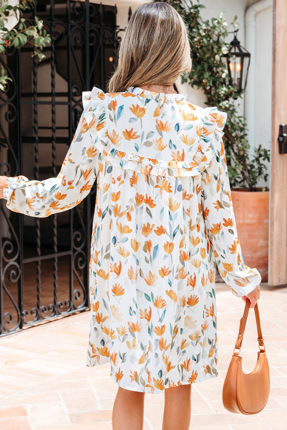 Woman wearing long sleeve floral ruffle dress with orange and yellow flowers, holding tan handbag