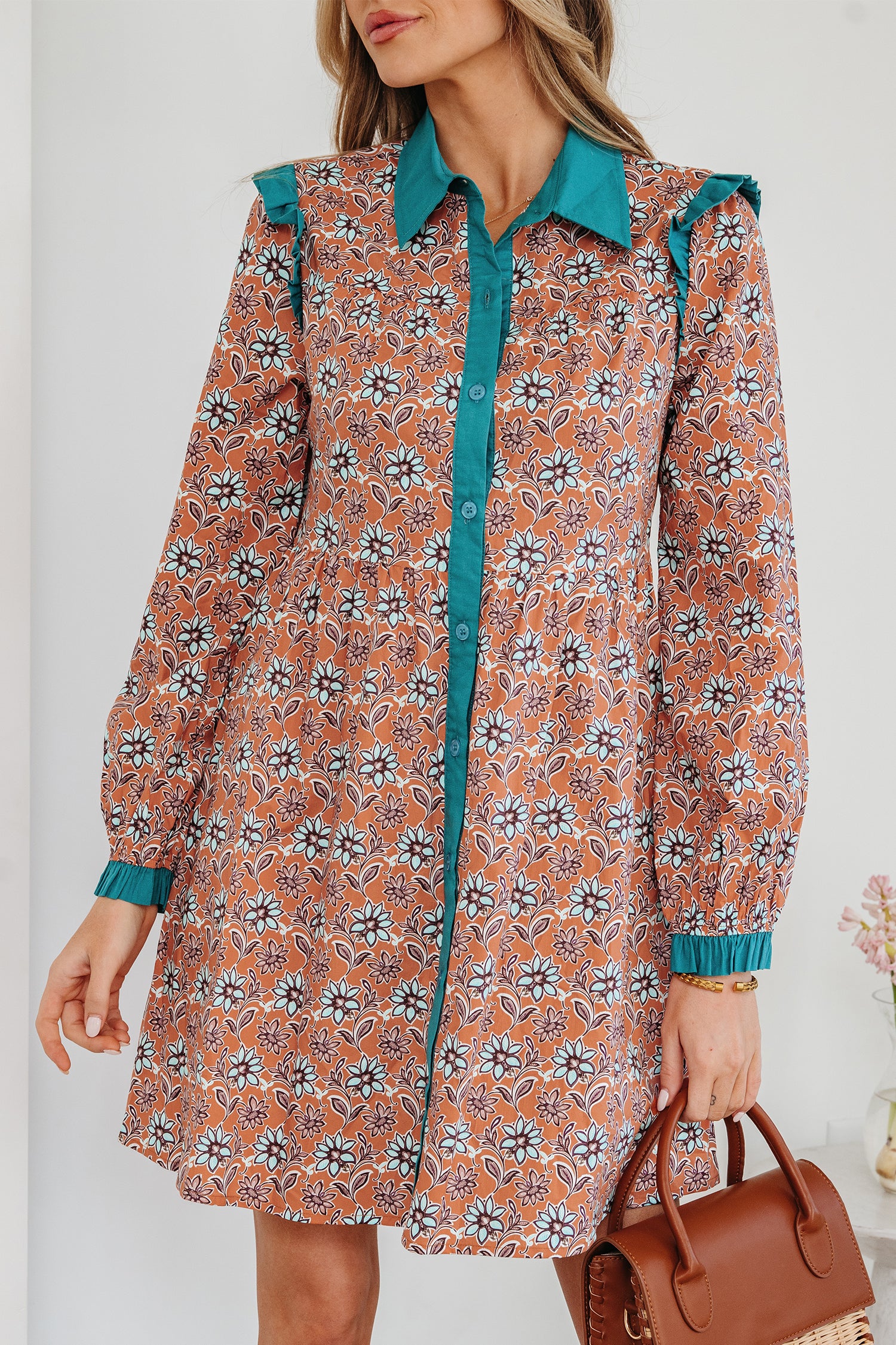 Woman wearing long-sleeve floral print shirt dress with teal collar and trim, holding a brown handbag
