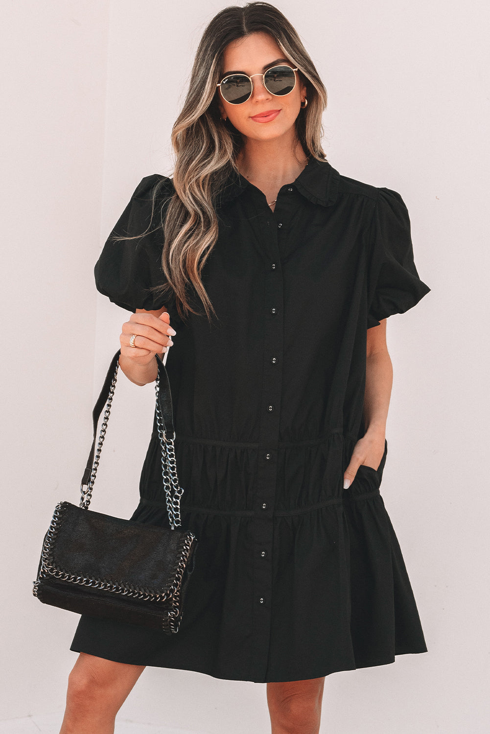 Woman wearing black puff sleeve button-up dress, round sunglasses, and holding a black chain shoulder bag