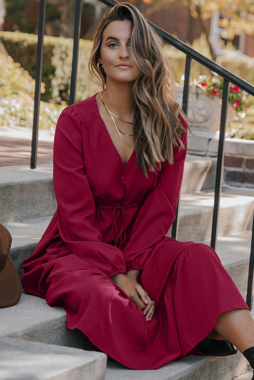 Woman in a long-sleeve red dress sitting on outdoor steps, wearing layered gold necklaces.