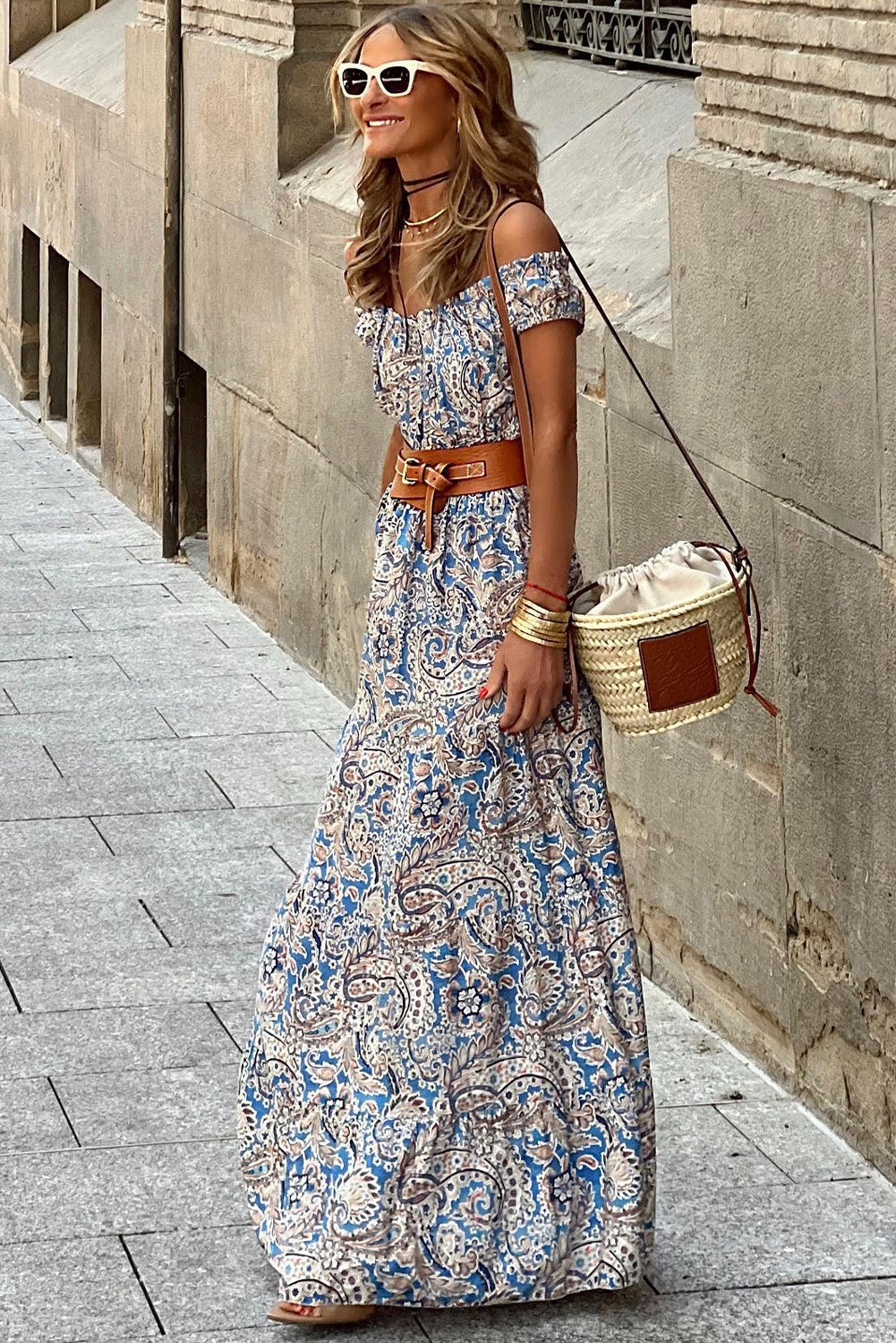 Woman wearing off-shoulder paisley maxi dress, brown belt, and straw crossbody bag on city street