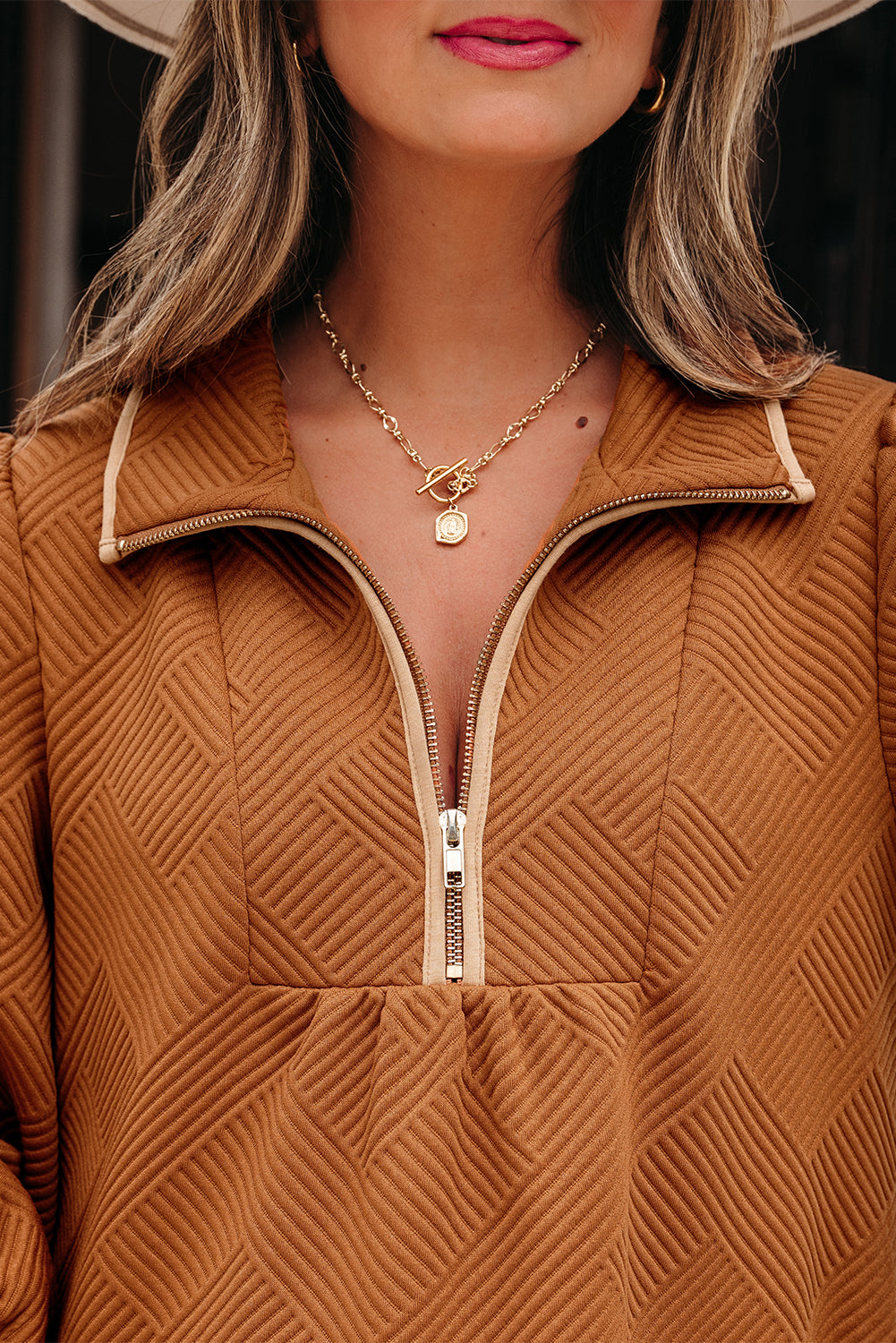 Woman wearing textured brown zip-up top with gold chain necklace and pink lipstick