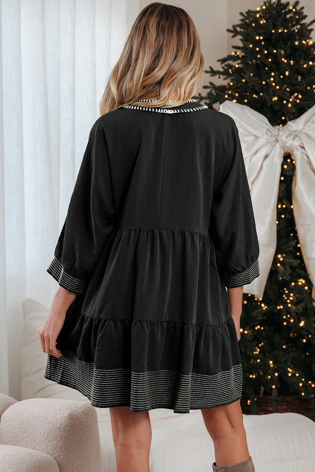 Woman in black long sleeve dress with striped trim, standing by decorated Christmas tree