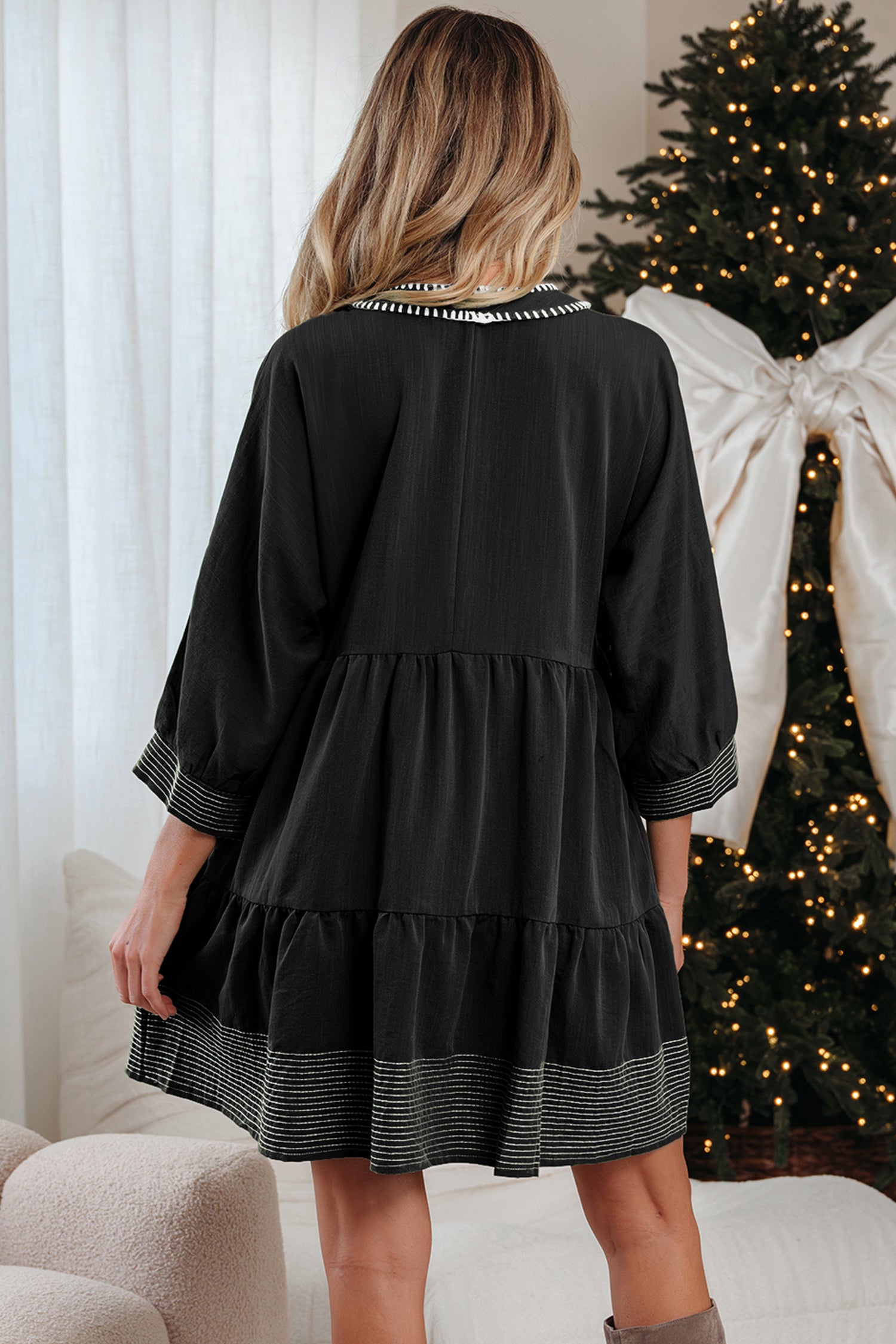 Woman in black long sleeve dress with striped trim, standing by decorated Christmas tree