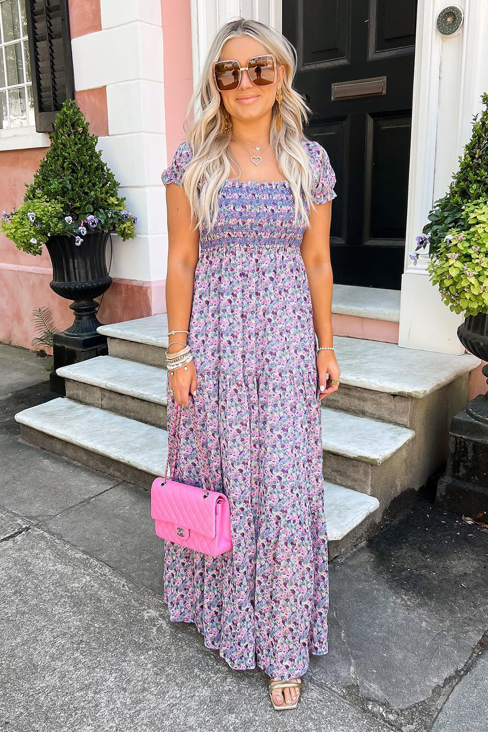 Woman in a floral maxi dress with pink quilted handbag, sunglasses, and sandals outside a home