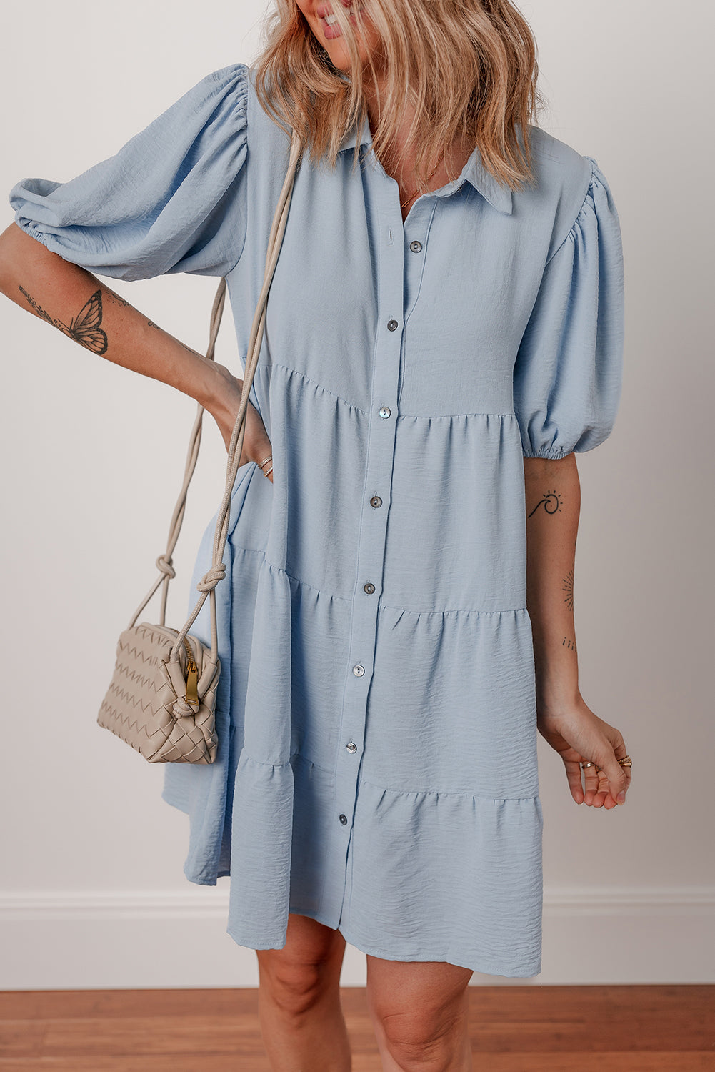 Woman in light blue button-up tiered mini dress with puff sleeves and beige woven handbag