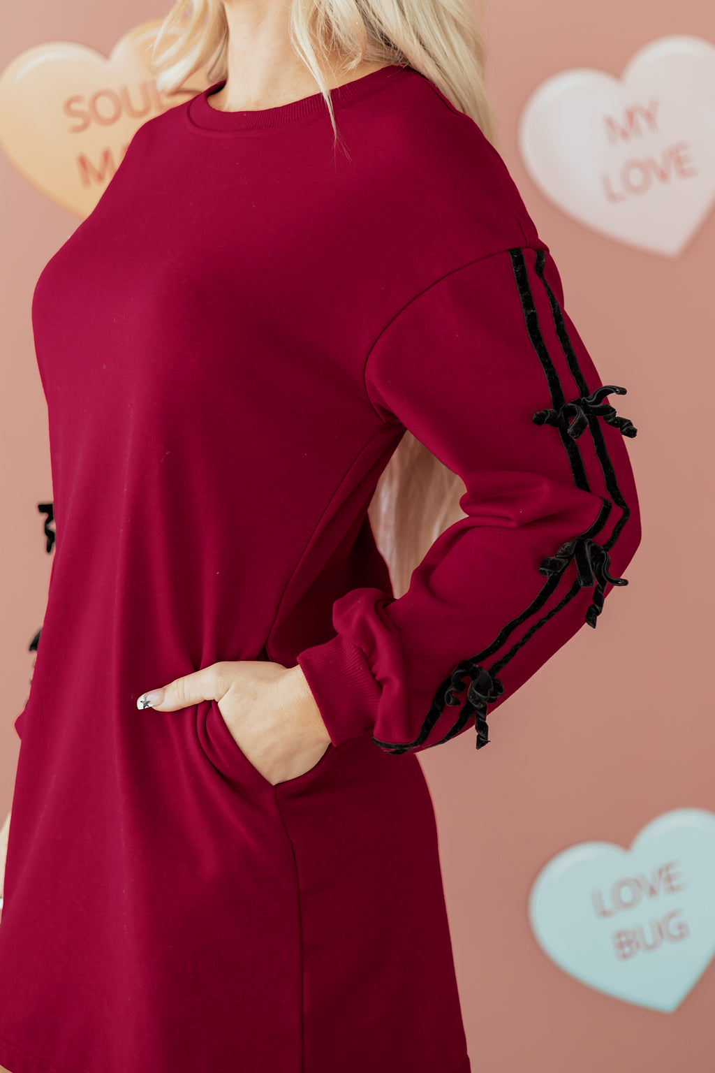 Woman in long sleeve burgundy dress with black ribbon accents, hand in pocket, pastel heart background