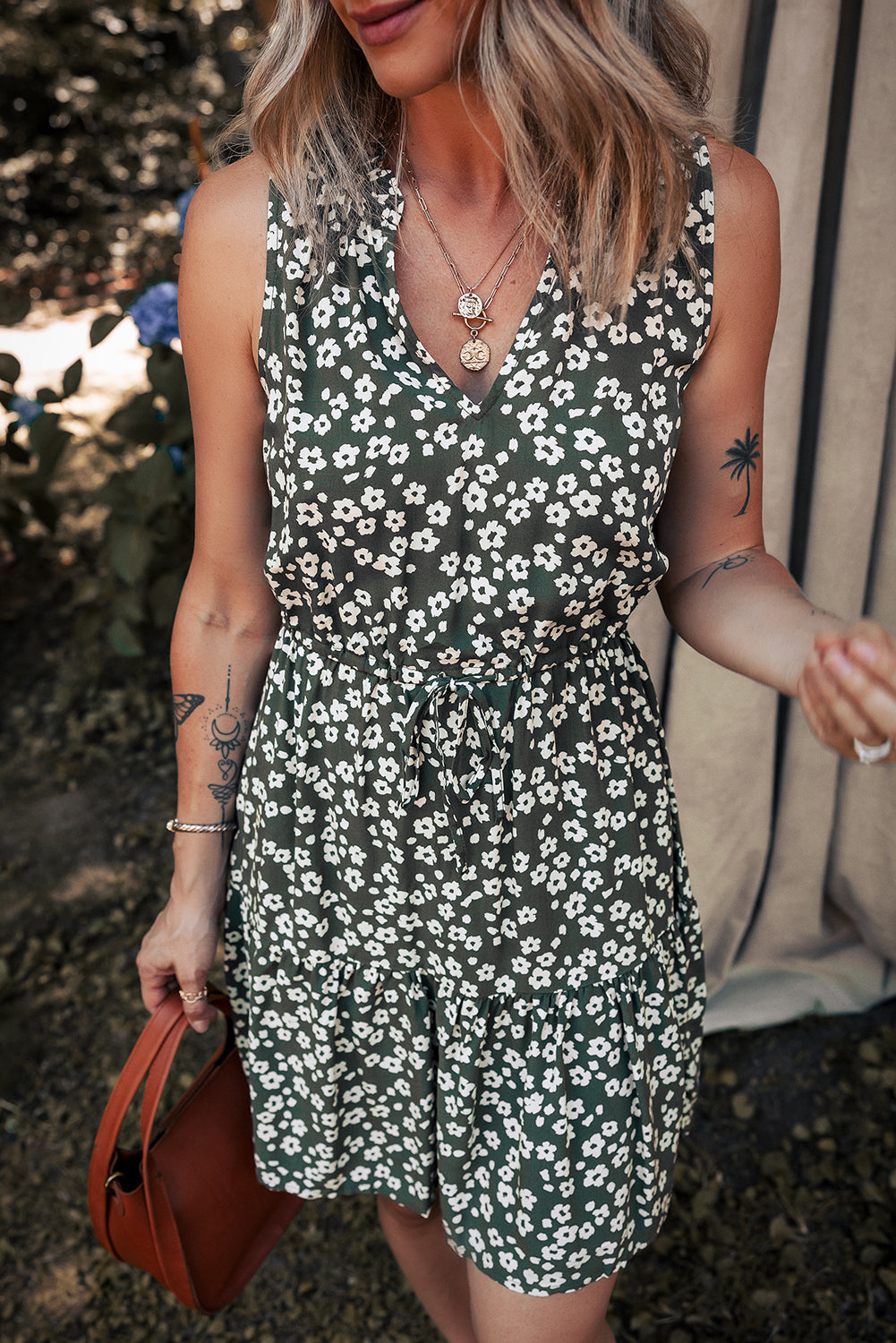 Woman in a sleeveless green floral dress holding a brown handbag, outdoor setting