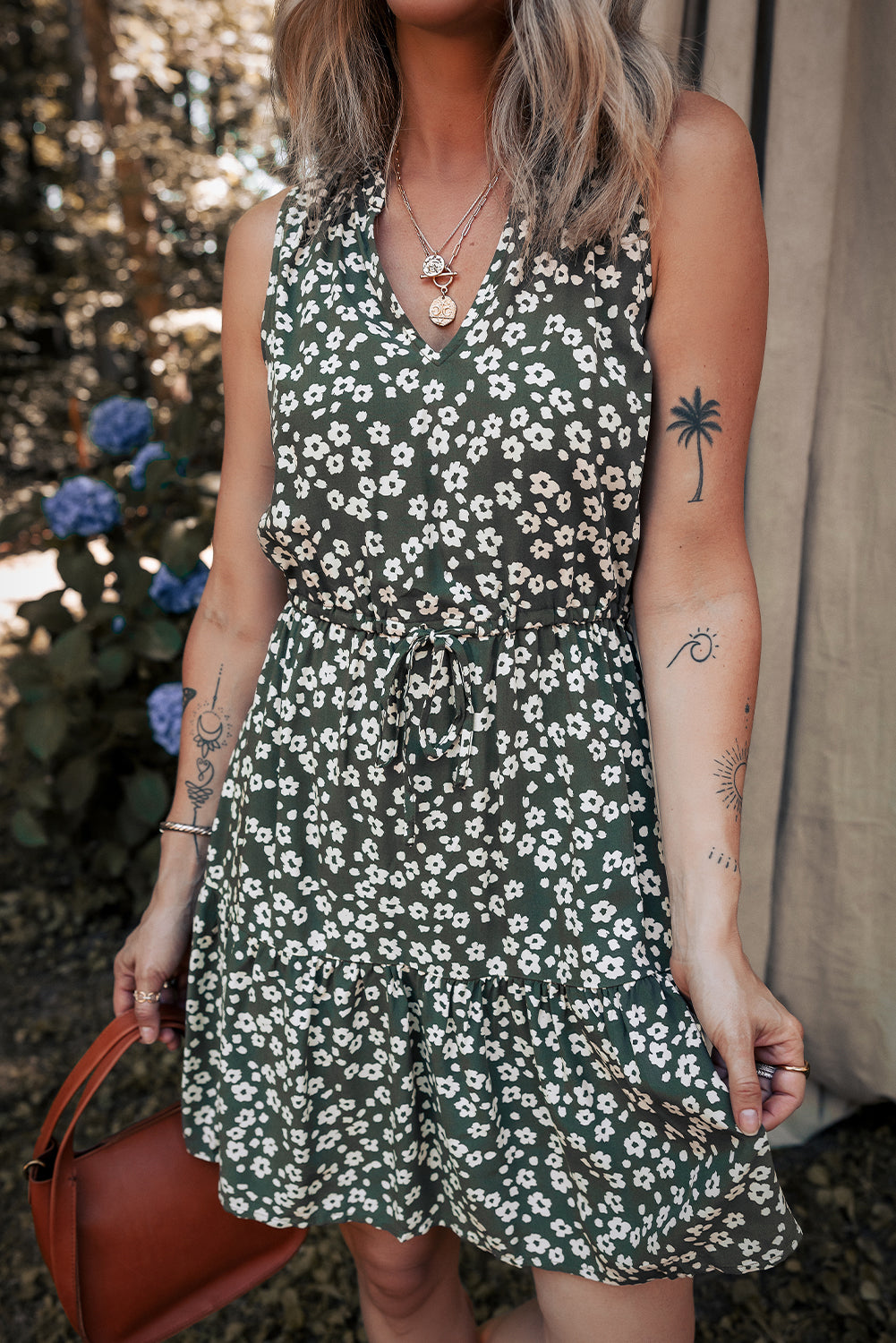 Woman in green floral sleeveless dress with drawstring waist, holding brown handbag outdoors