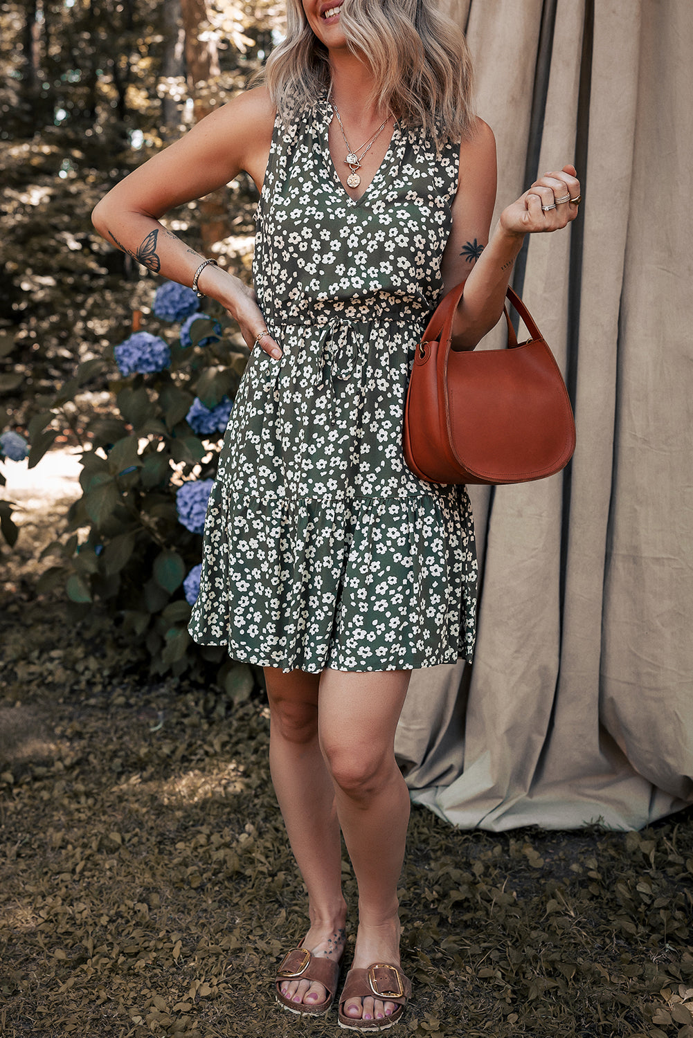 Woman in green floral sleeveless dress holding brown handbag, standing outdoors