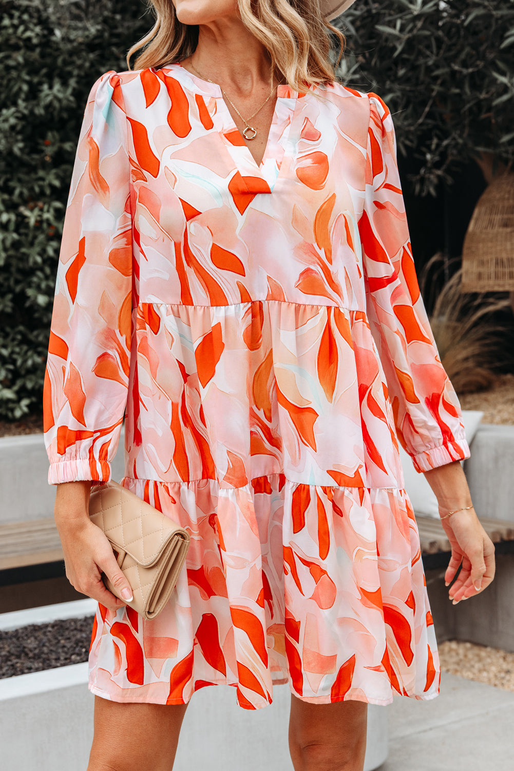 Woman wearing a flowy long sleeve dress with orange and pink abstract floral print, holding a beige clutch
