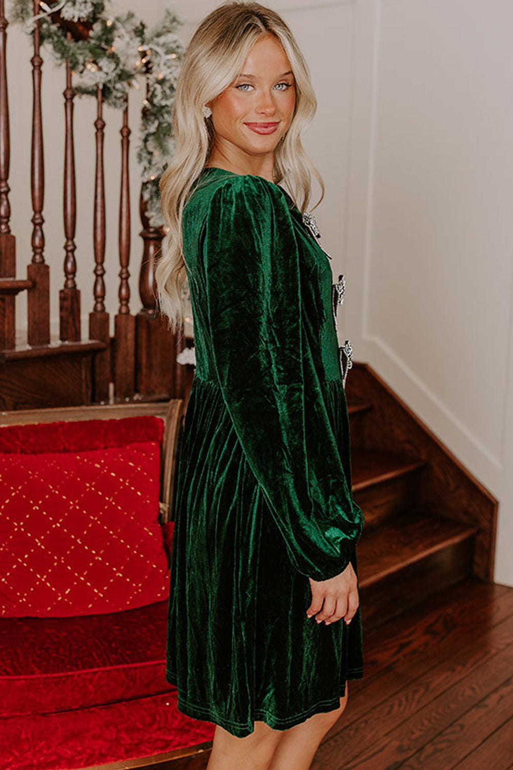 Woman in green velvet long-sleeve dress standing by staircase with holiday decor