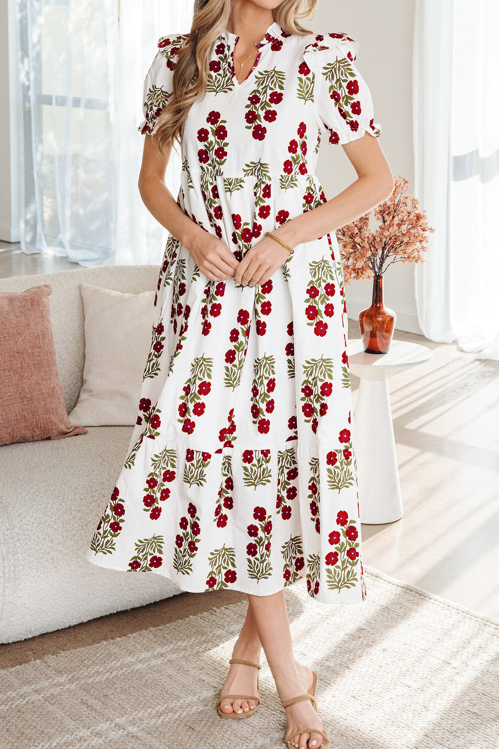 Woman wearing a white floral midi dress with red flowers and green leaves, standing in a bright living room