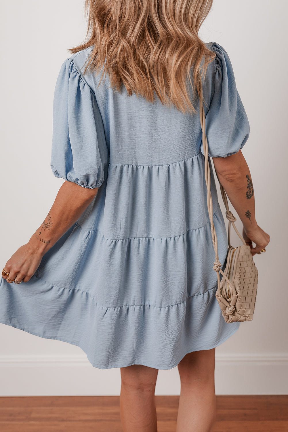 Woman wearing a light blue tiered short dress with puff sleeves and a beige woven crossbody bag