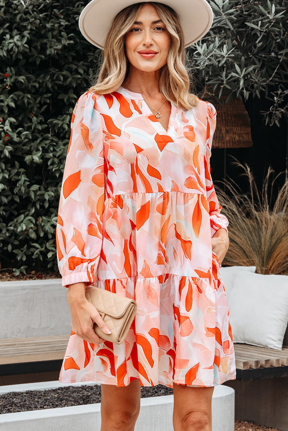 Woman in a colorful abstract print dress with long sleeves and a hat, holding a beige clutch