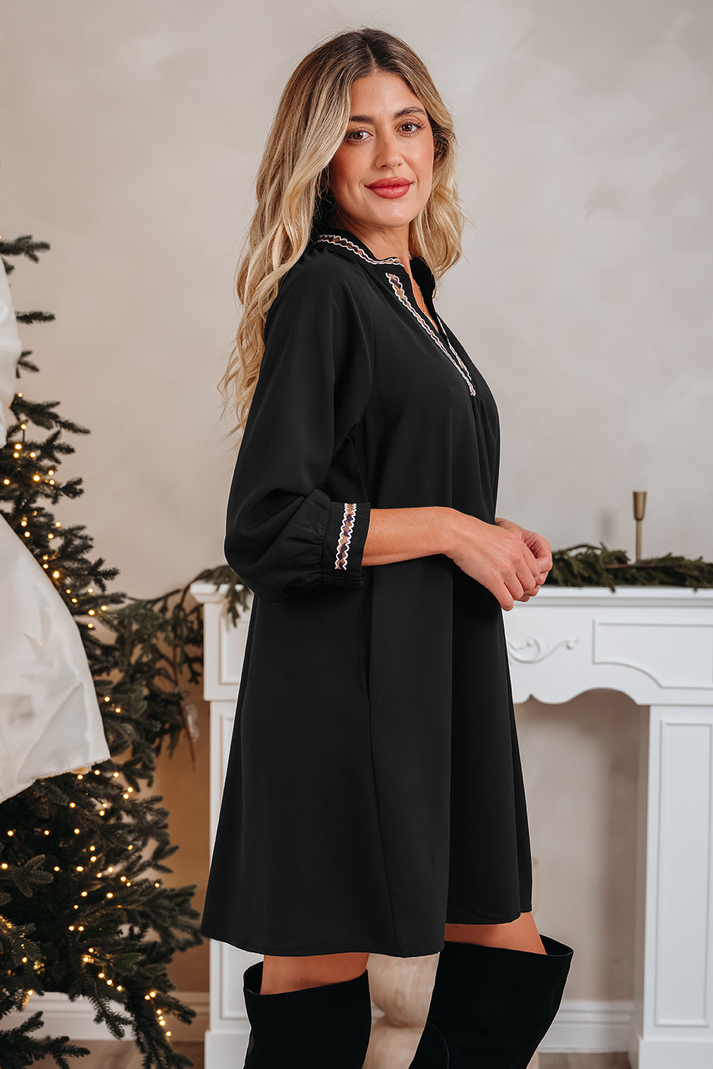 Woman in black long-sleeve tunic dress with embroidered trim, standing indoors