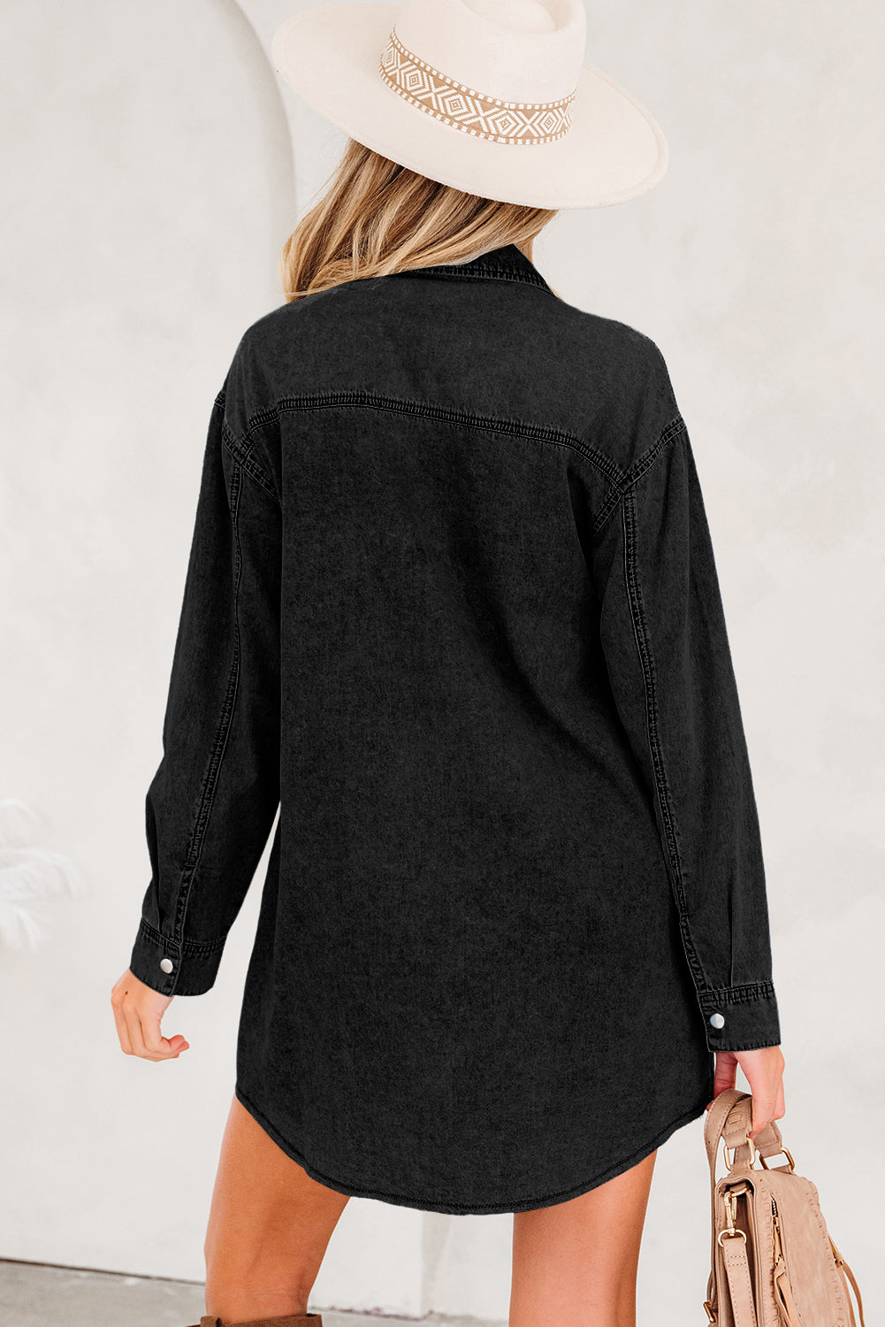 Woman in black denim shirt dress and beige hat, holding a tan handbag, back view