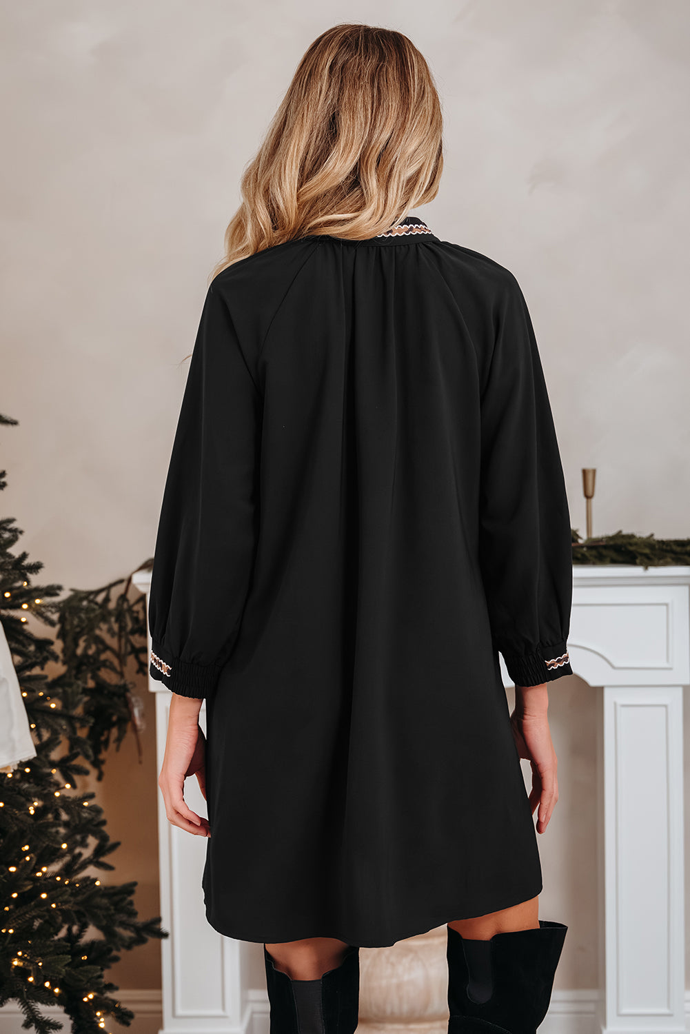 Woman in a black long sleeve dress and boots, standing indoors by a decorated Christmas tree