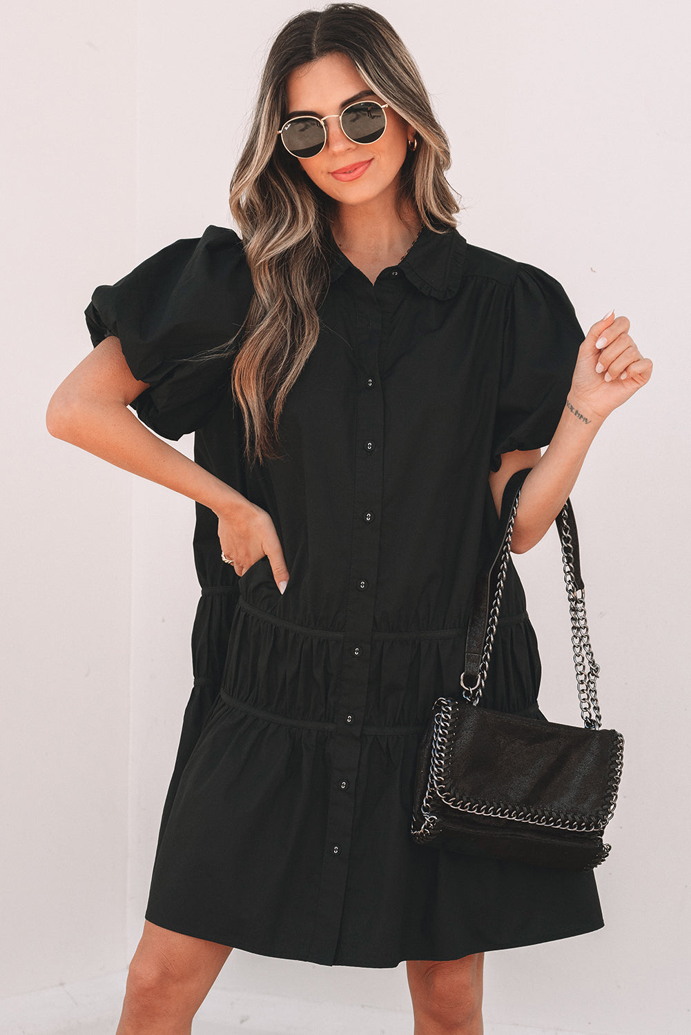 Woman in black button-down mini dress with puff sleeves, black handbag, and sunglasses