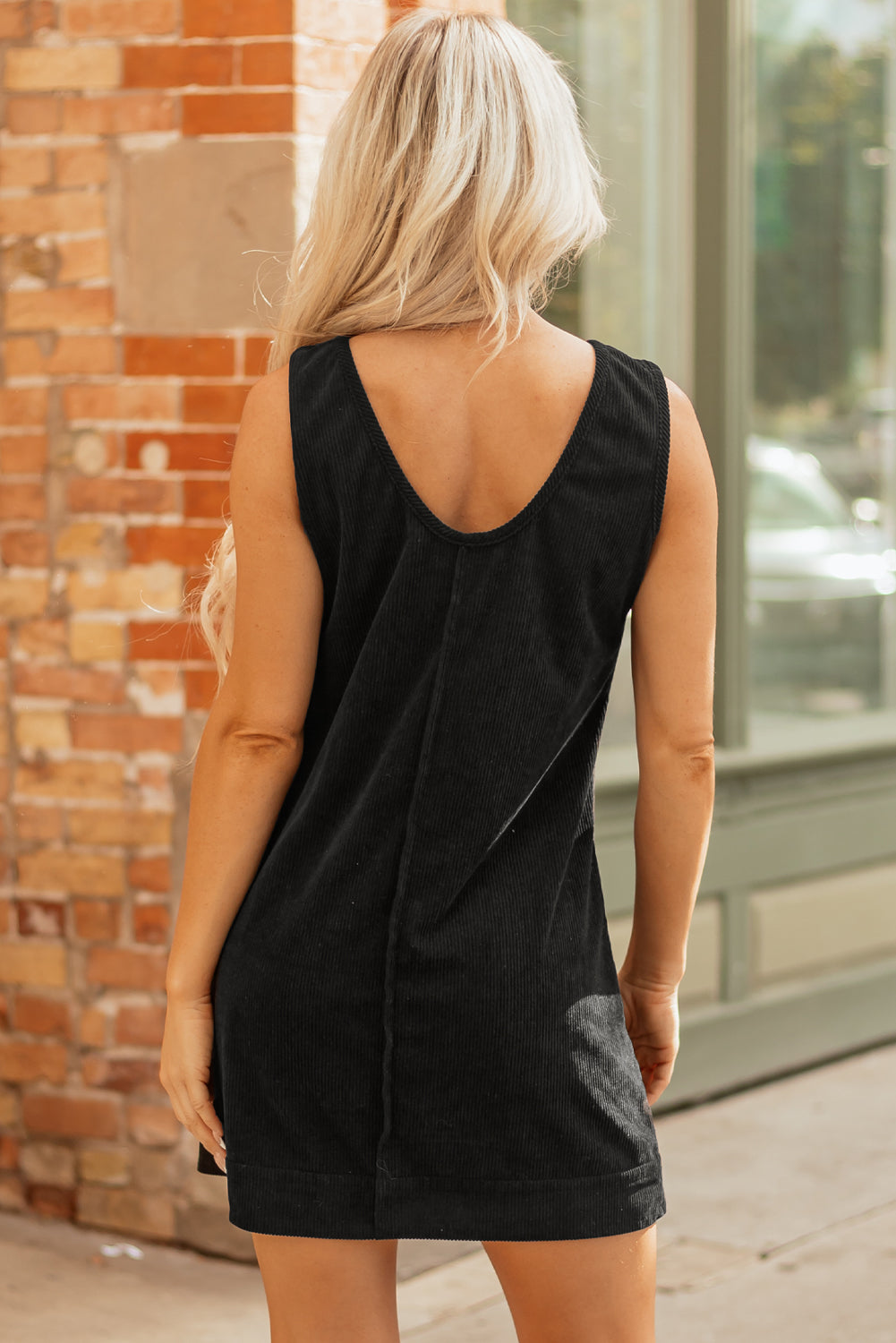 Woman in sleeveless black corduroy dress with V-back, standing by a brick wall outdoors