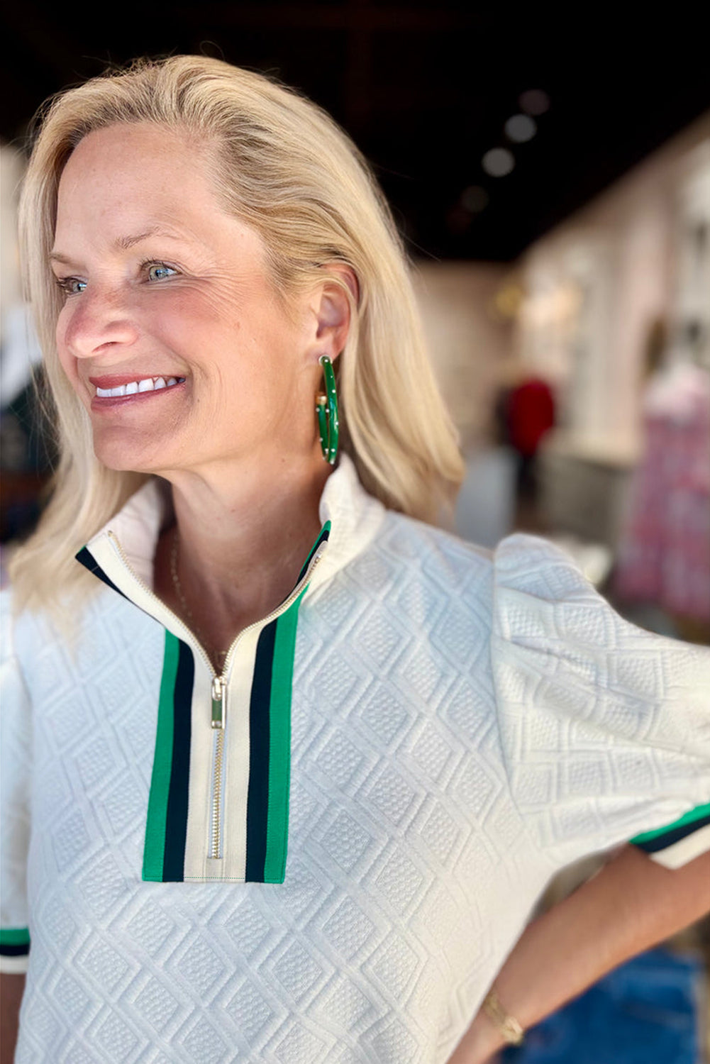 Woman wearing white textured short-sleeve top with green striped zip collar, smiling indoors