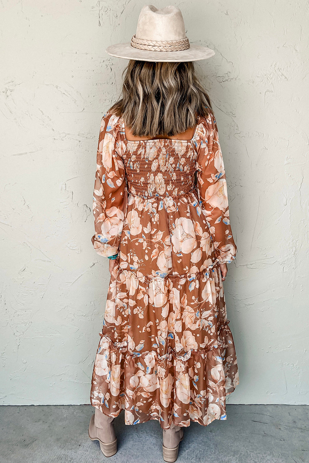 Woman in a brown floral maxi dress, wide-brim hat, and boots, standing against a textured wall