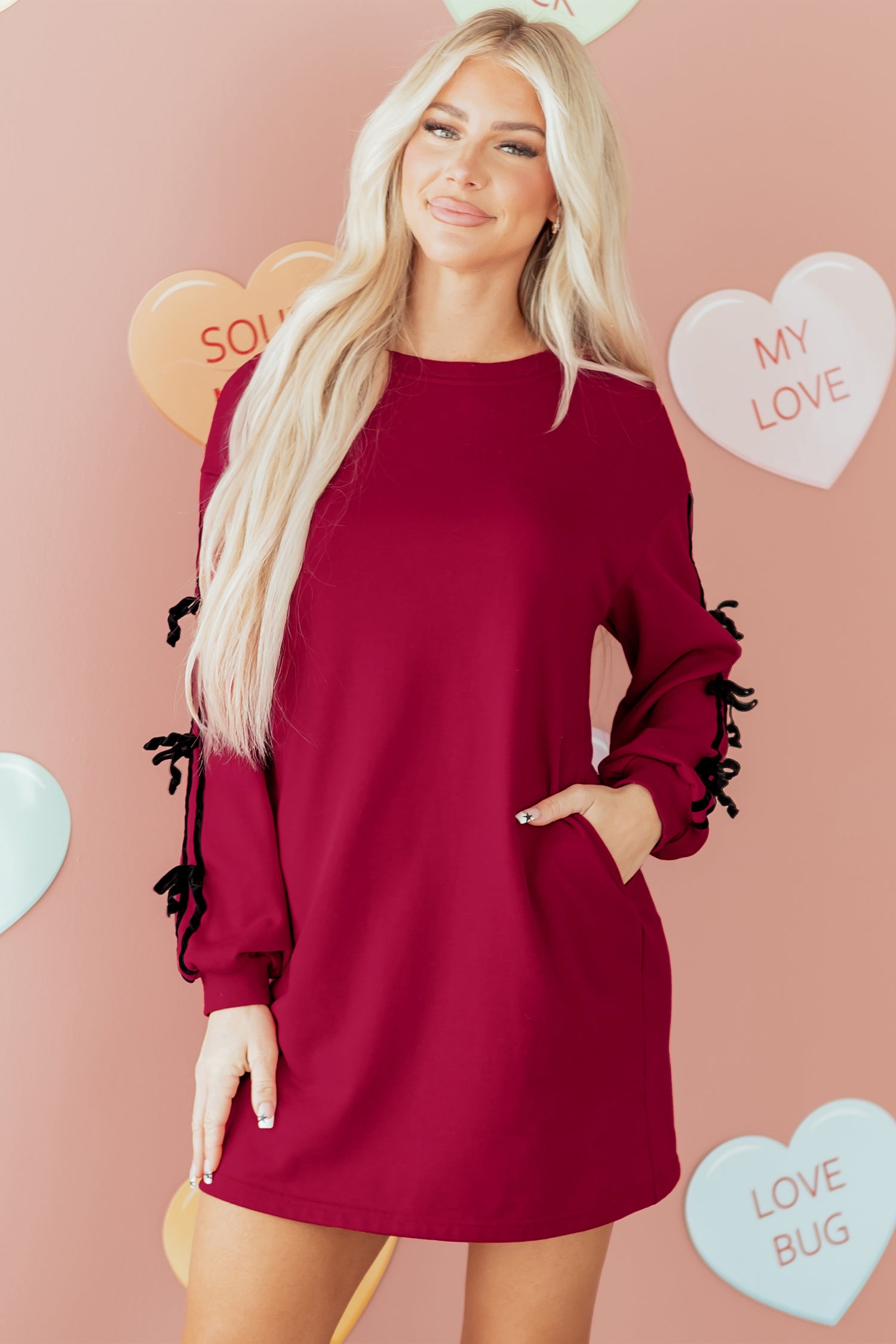 Woman in a long sleeve burgundy dress with side bows, standing by pastel heart decorations