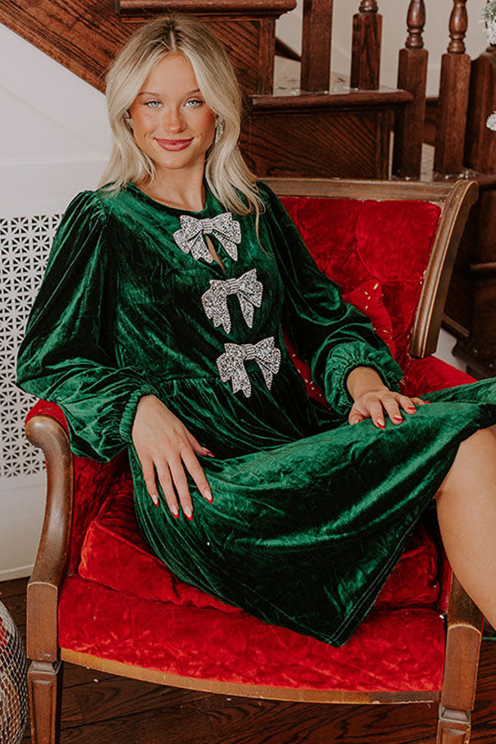 Woman in green velvet dress with rhinestone bow accents sitting on red velvet chair