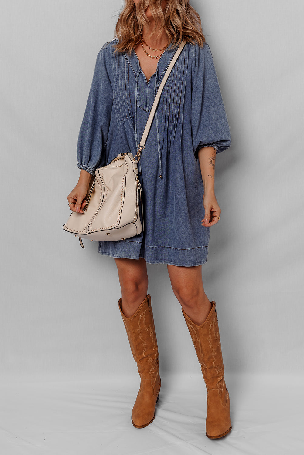 Woman in denim dress with beige shoulder bag and tan knee-high boots against gray background