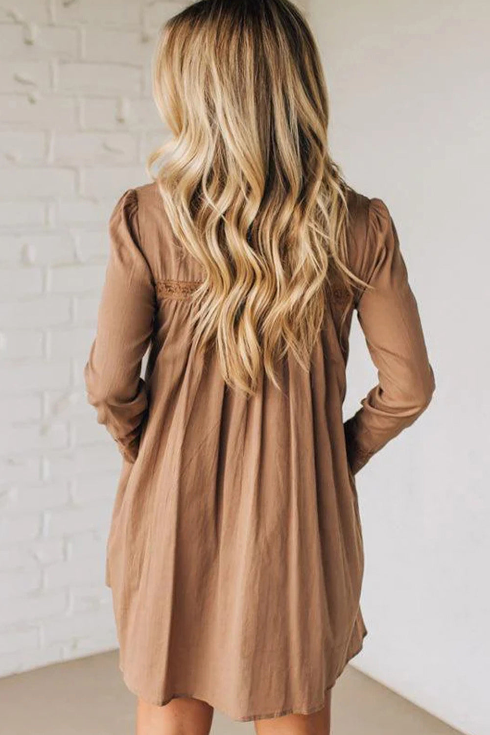 Woman with long wavy blonde hair wearing loose brown long sleeve dress, back view